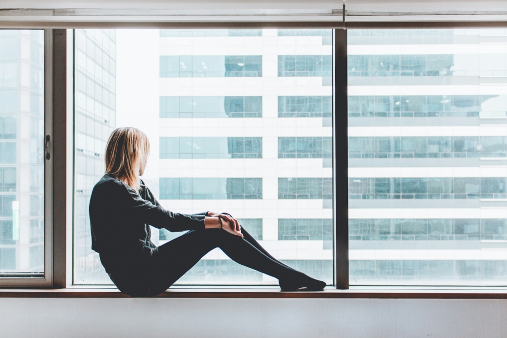 Een vrouw zit op een vensterbank en kijkt naar buiten door een groot raam met dubbel glas. Ze draagt een donkere trui en legging en lijkt diep in gedachten verzonken terwijl ze naar de hoge gebouwen aan de overkant van de straat kijkt. Het dubbel glas biedt een helder uitzicht en isoleert de kamer van het geluid en de kou van buiten.