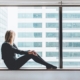 Een vrouw zit op een vensterbank en kijkt naar buiten door een groot raam met dubbel glas. Ze draagt een donkere trui en legging en lijkt diep in gedachten verzonken terwijl ze naar de hoge gebouwen aan de overkant van de straat kijkt. Het dubbel glas biedt een helder uitzicht en isoleert de kamer van het geluid en de kou van buiten.